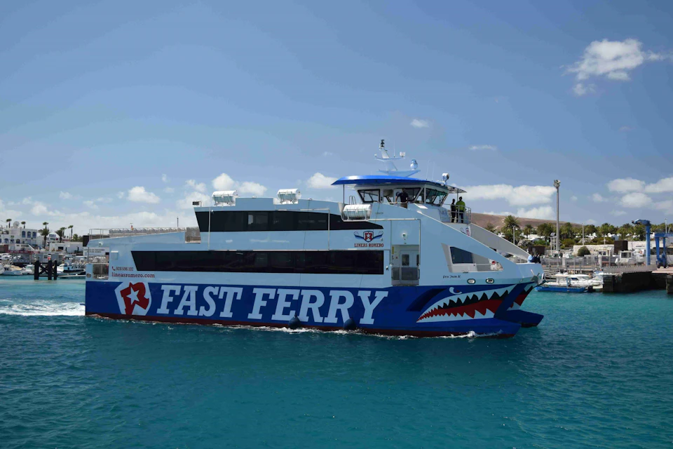 A blue and white ferry labeled "FAST FERRY" with shark graphics sails on clear blue water under a partly cloudy sky.