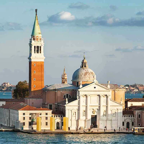 Ett historiskt strandnära komplex med en stor kyrka, högt klocktorn och intilliggande byggnader, omgivna av vatten med båtar vid bryggan.
