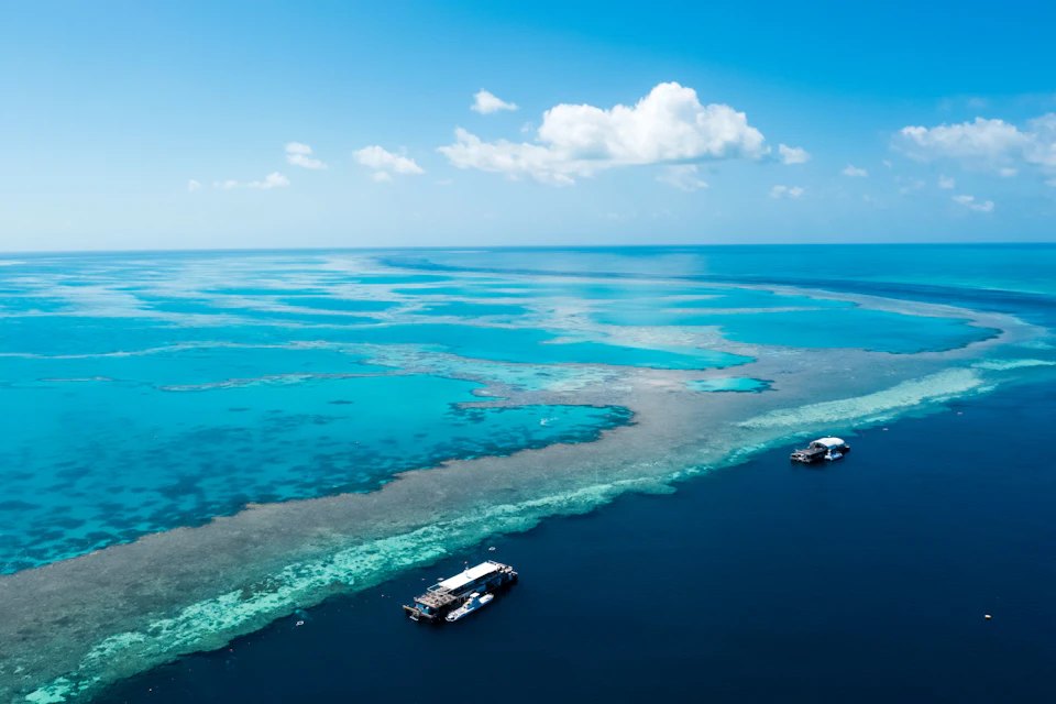 Two boats are anchored near a coral reef in clear, turquoise water under a bright blue sky with a few scattered clouds.