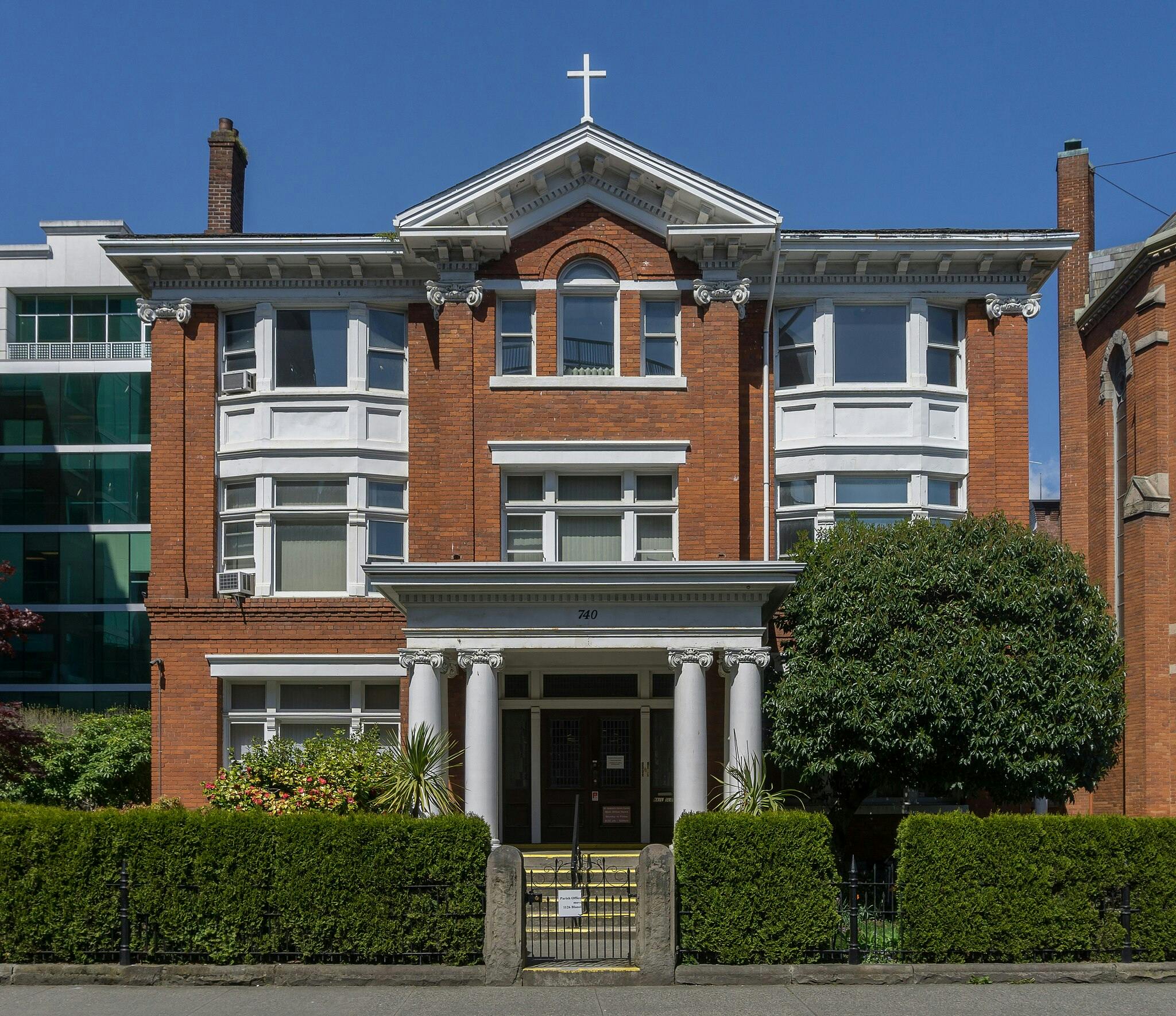 Three-story red-brick building with white-trimmed windows and columns, a cross on top, and a gated entrance surrounded by greenery.