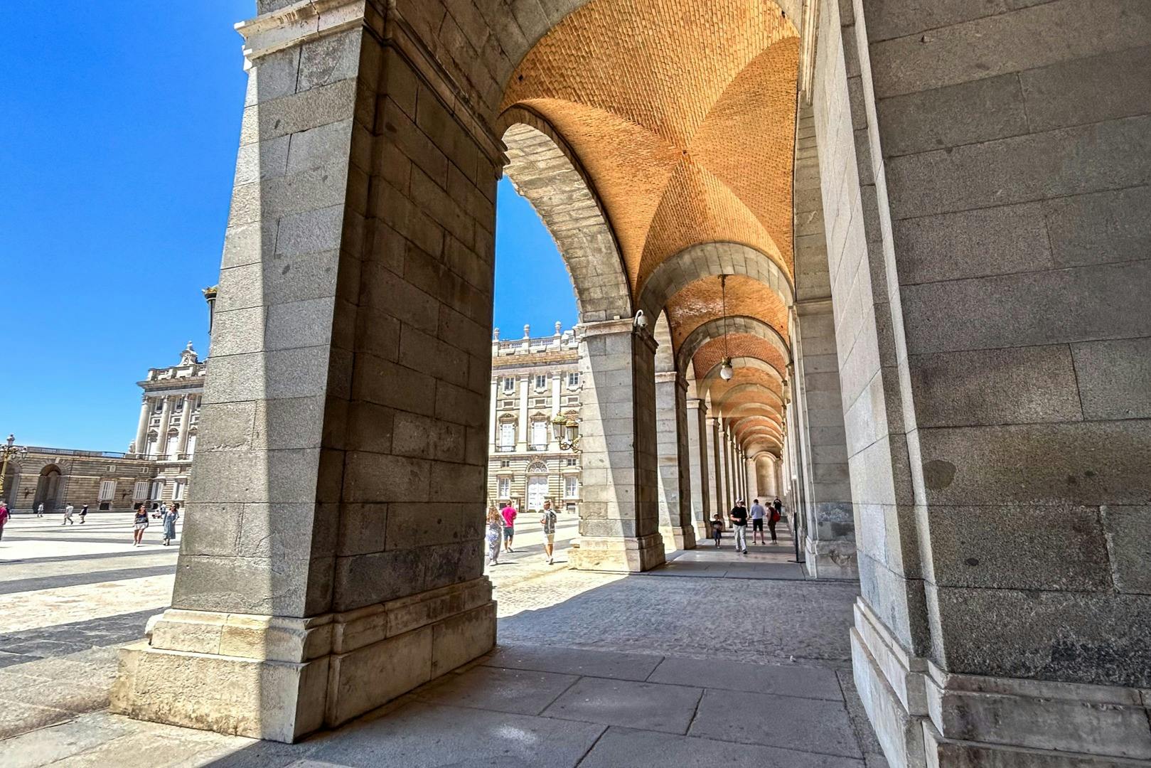 Arco de pedra com um teto abobadado e pessoas a caminhar por baixo, com um edifício histórico visível ao fundo.