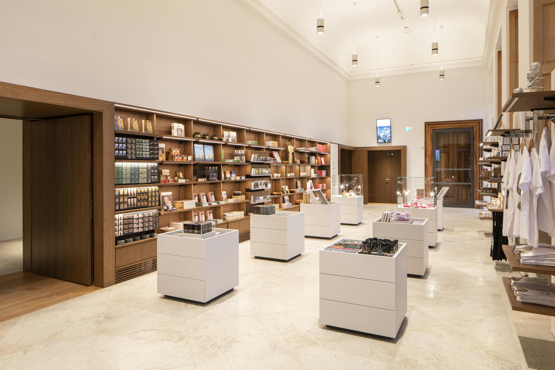 Ένας μοντέρνος εσωτερικός χώρος καταστήματος με ξύλινα ράφια επενδεδυμένα με προϊόντα, πολλά λευκά τραπέζια και κρεμαστά φωτιστικά από πάνω.