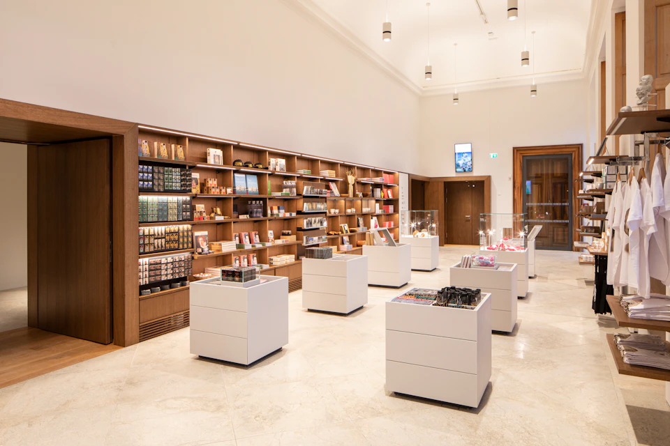 A modern store interior with wooden shelves lined with products, several white display tables, and pendant lights above.