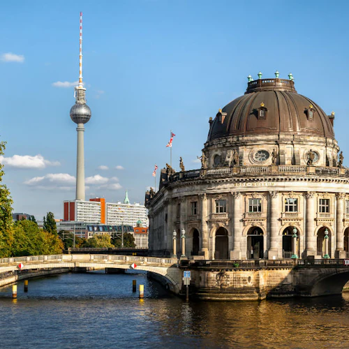 En välvd historisk byggnad vid en flod, med en bro och ett högt tv-torn i bakgrunden under en blå himmel.