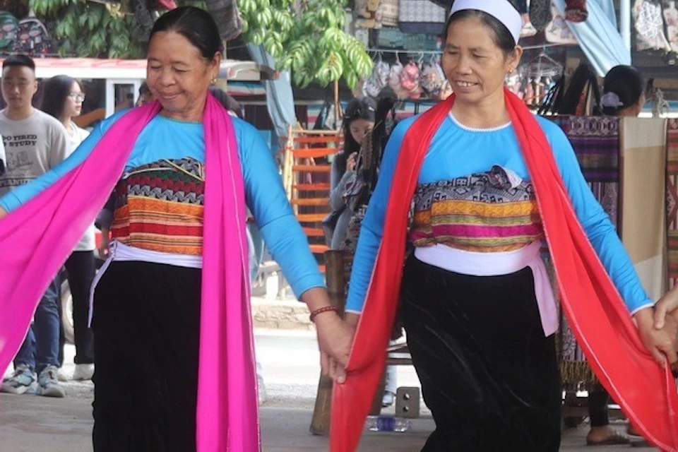 Three women in colorful traditional attire perform a folk dance under a metal roof, surrounded by spectators and market stalls.