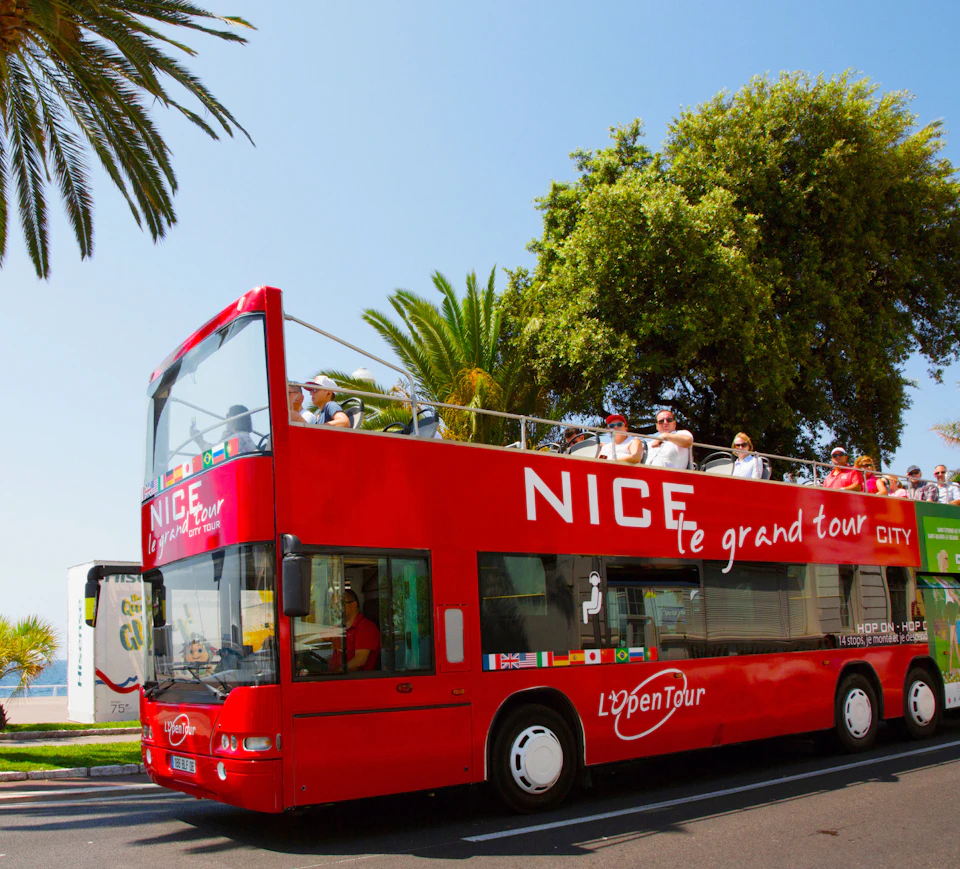 A red double-decker tour bus labeled "Nice, le grand tour" with passengers on the open-top deck, parked near palm trees.