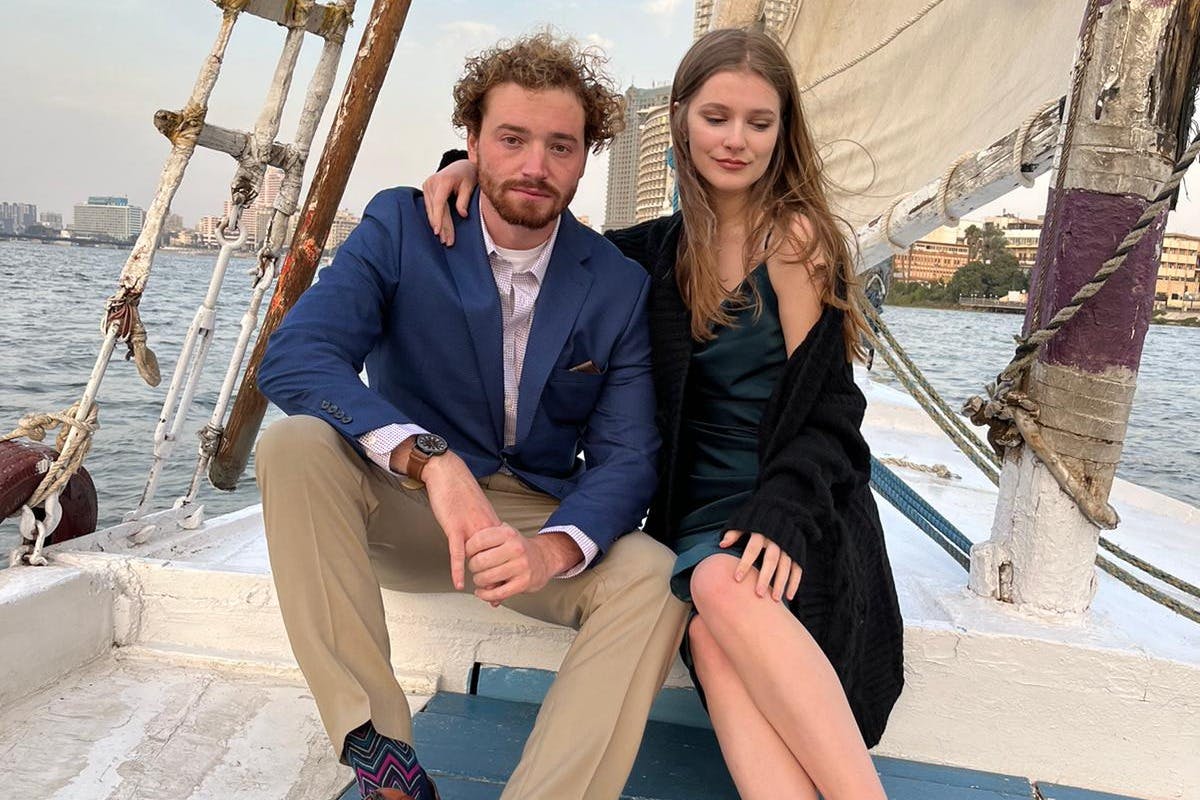 一男一女坐在船上，背景是风帆和城市景观；男士穿着蓝色西装外套，女士穿着深色连衣裙和黑色开衫。