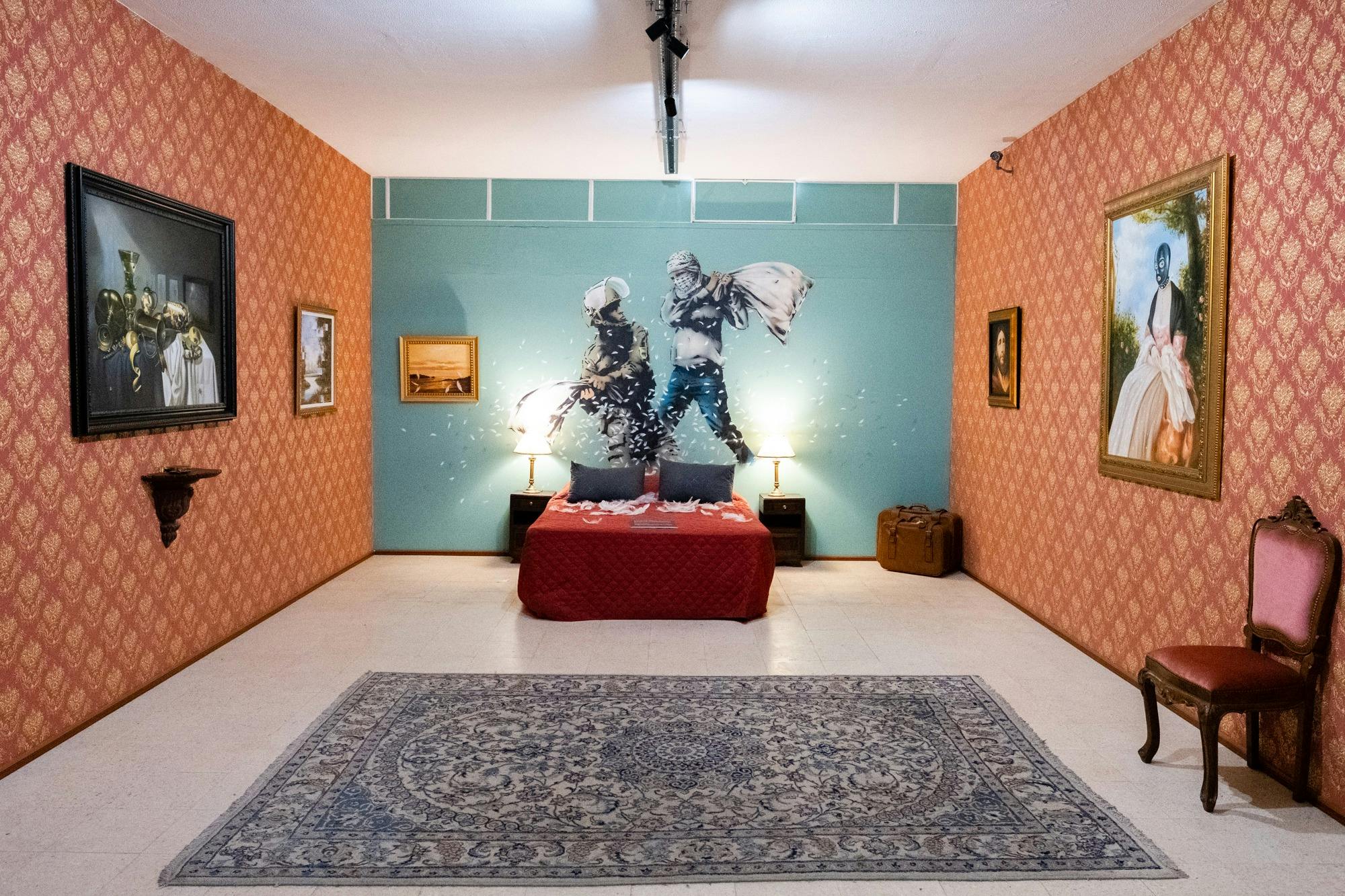 Υπνοδωμάτιο με κόκκινο κάλυμμα κρεβατιού, μαξιλάρια και μπλε τοίχο με γκράφιτι δύο ατόμων που χορεύουν. Κορνιζαρισμένες εικόνες στους τοίχους.