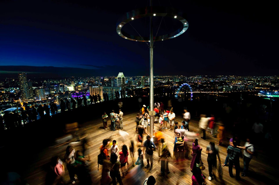 Crowds of people gather on an observation deck at night overlooking a brightly lit city skyline.