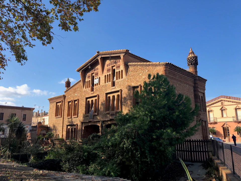 A large brick building with decorative chimneys, surrounded by greenery and partially obscured by trees, under a clear blue sky.