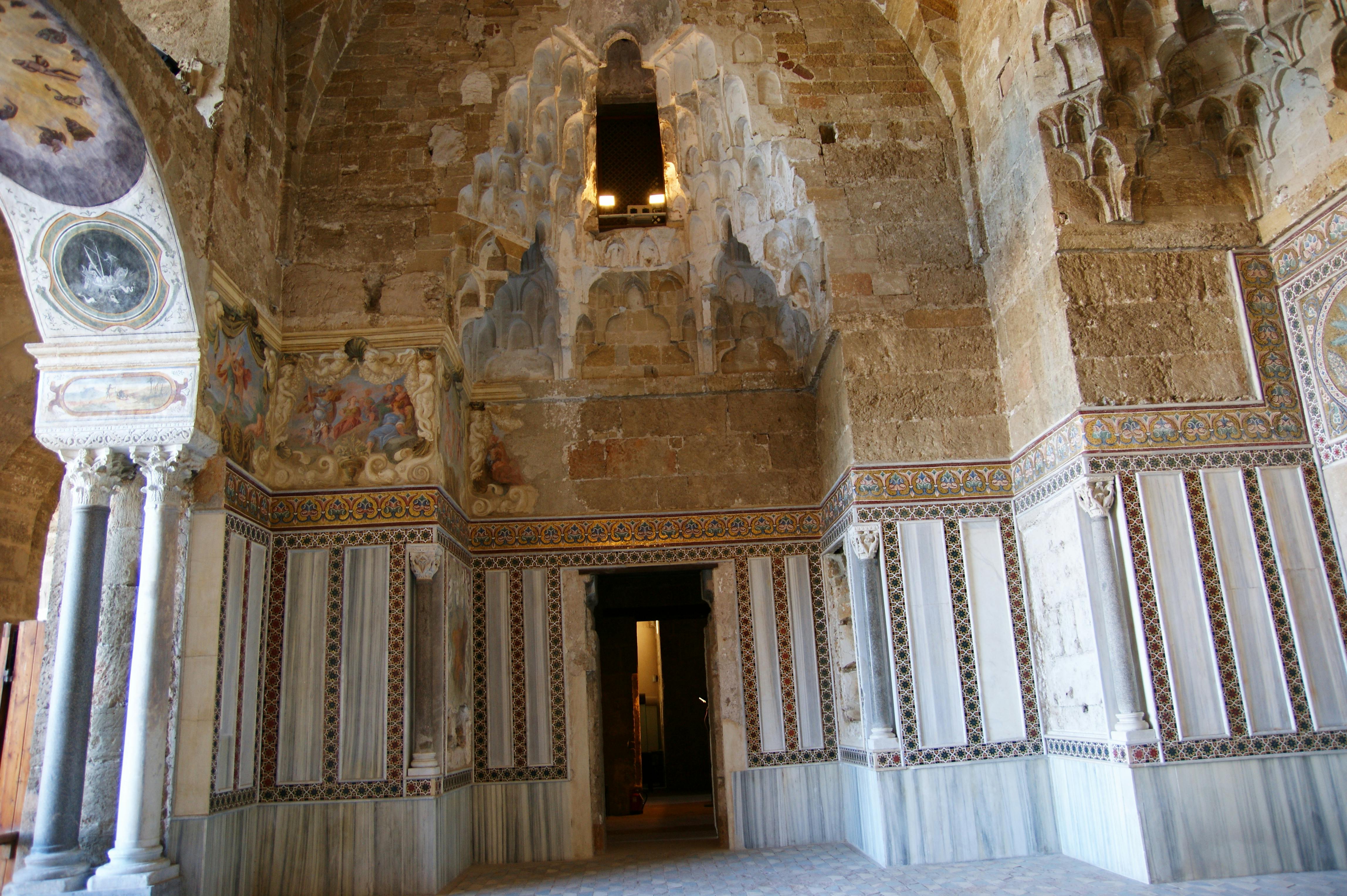 Interior of an ornate, historical stone building with decorative arches, patterned trim, and wall frescoes.