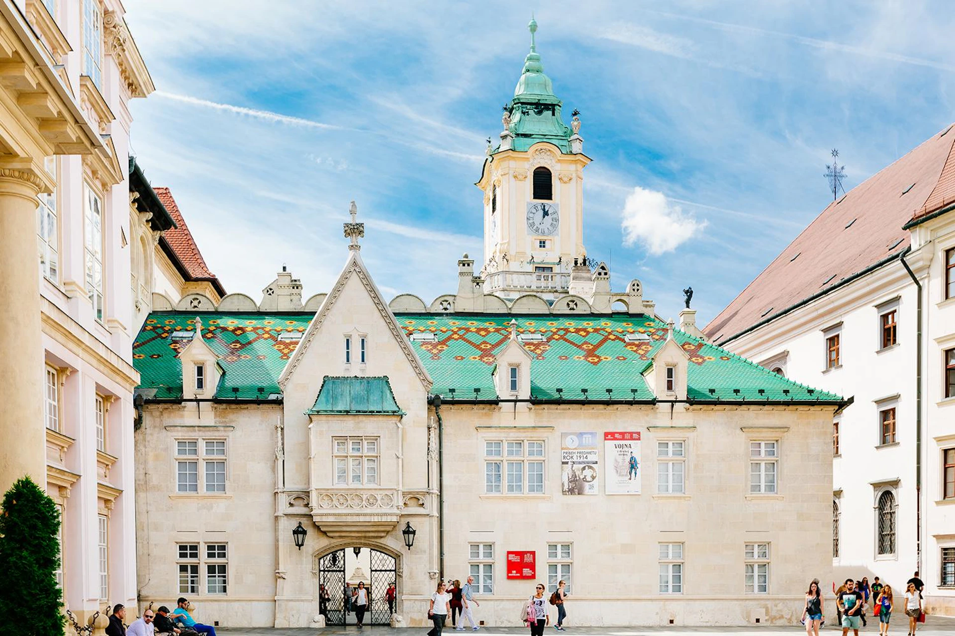 Historic building with a green tiled roof and clock tower, people walking in front, and posters on the façade under a blue sky.