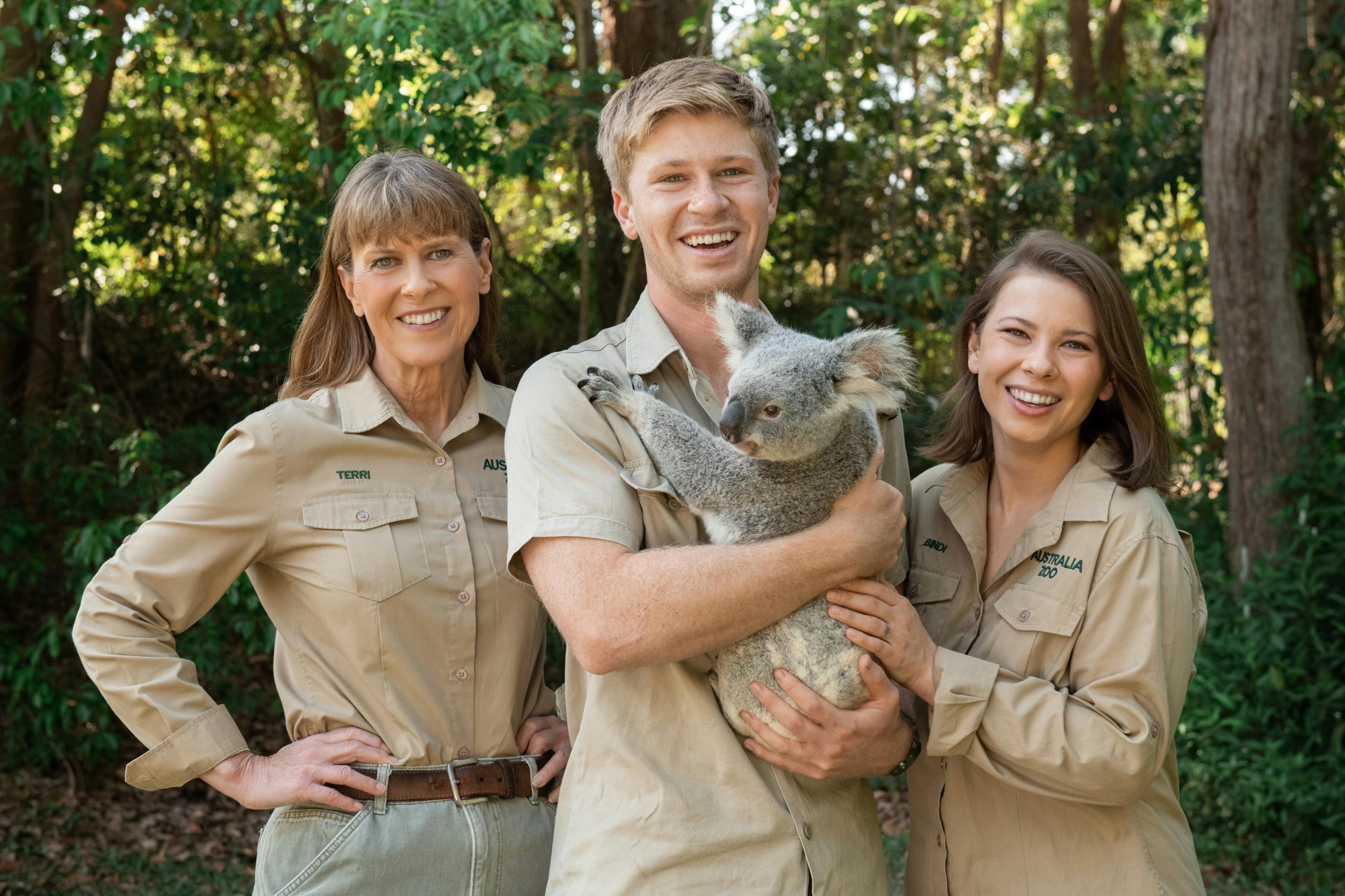 Drei lächelnde Personen in khakifarbenen Uniformen mit einem Koala, die im Freien vor einer üppigen Grünanlage stehen.