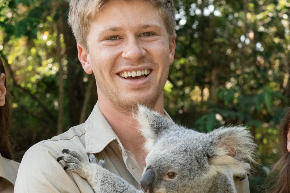 Three smiling individuals in khaki uniforms with a koala, standing outdoors in front of lush greenery.