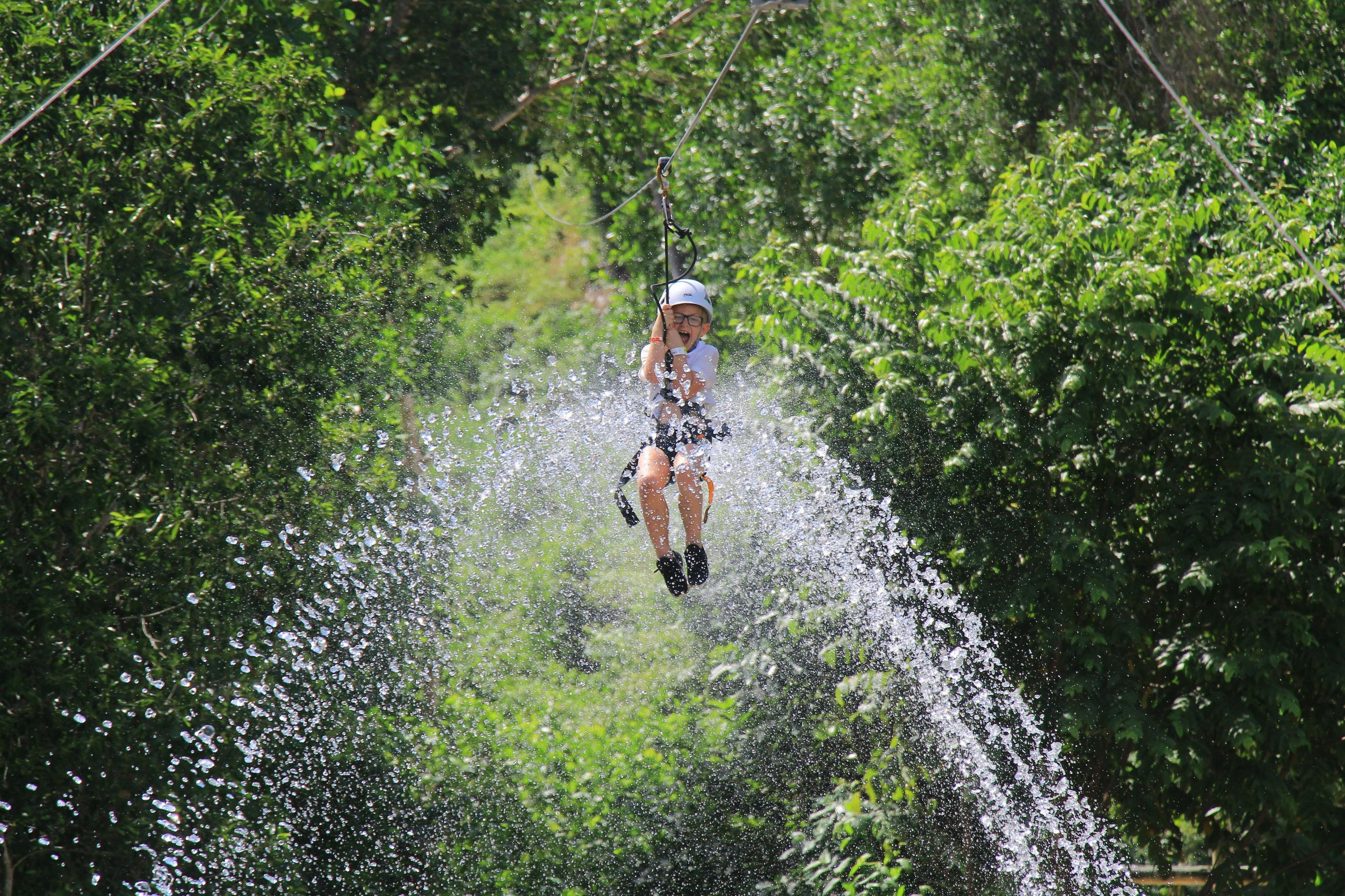 A person ziplining through a forested area with water splashing around them.