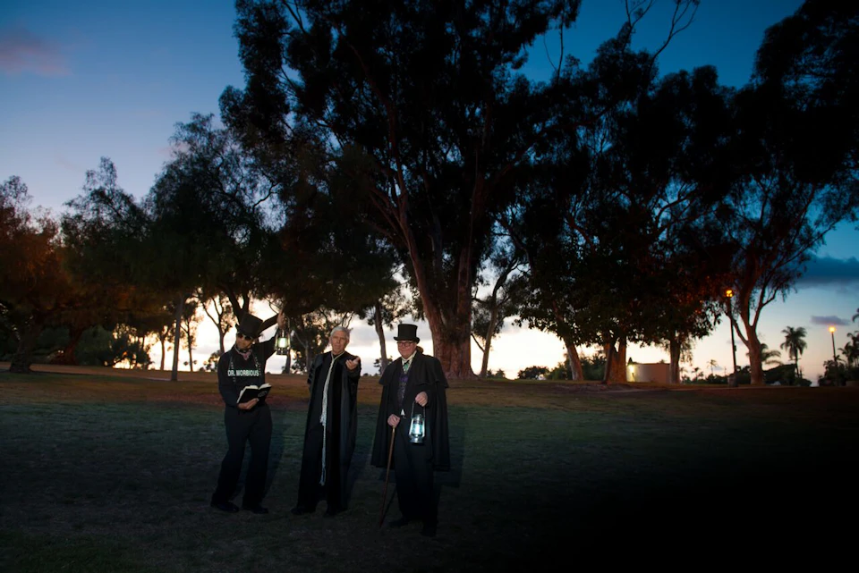 Three people with lanterns and dark clothing stand on grass near trees at dusk. One holds a book and wears a hat labeled "Old Morgue."