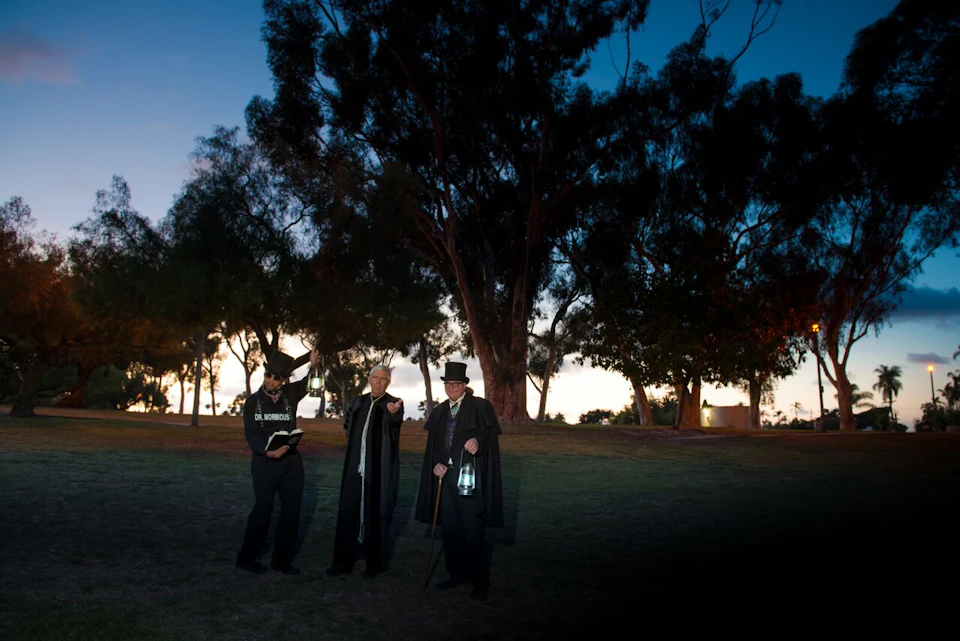 Three people with lanterns and dark clothing stand on grass near trees at dusk. One holds a book and wears a hat labeled "Old Morgue."