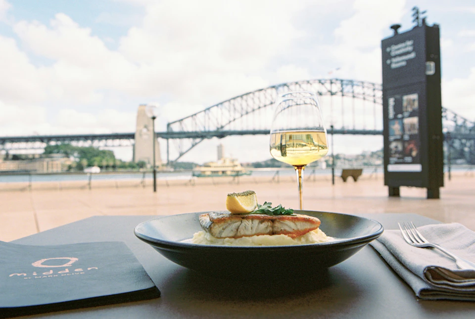 A plate with fish and mashed potatoes, a glass of white wine, and a bridge in the background under a partly cloudy sky.