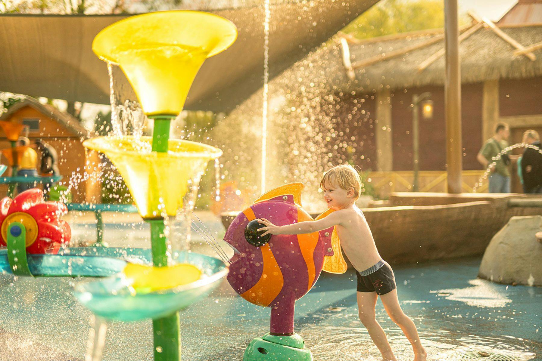 A child plays with a colorful fish-shaped water feature in a sunny splash park with thatched-roof structures in the background.