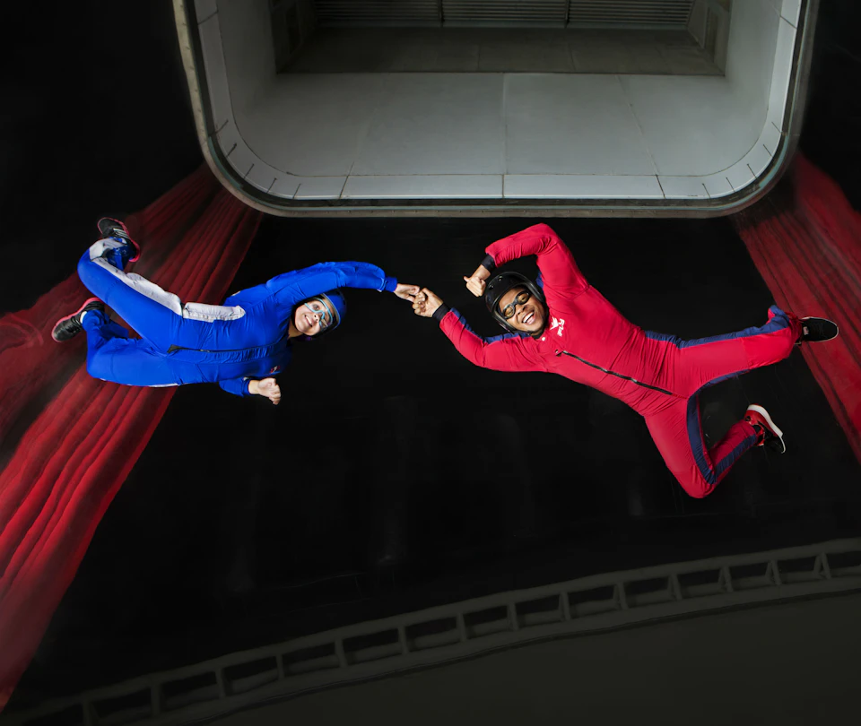 Two people in red and blue jumpsuits are indoor skydiving, floating against a background of red curtains and a white ceiling.