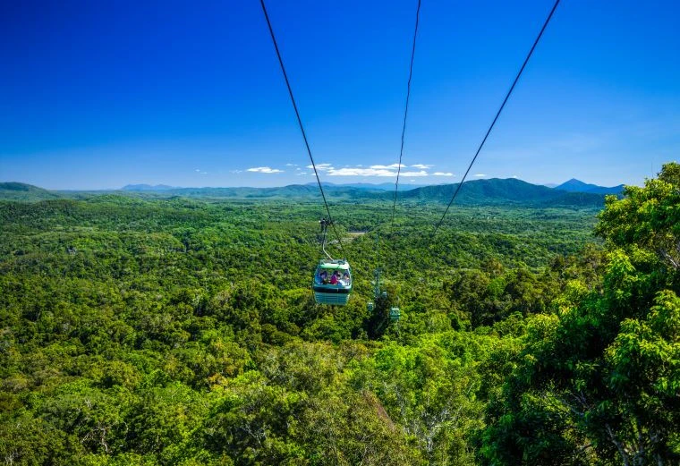 A cable car with passengers travels above a lush green forest with mountains in the background under a clear blue sky.