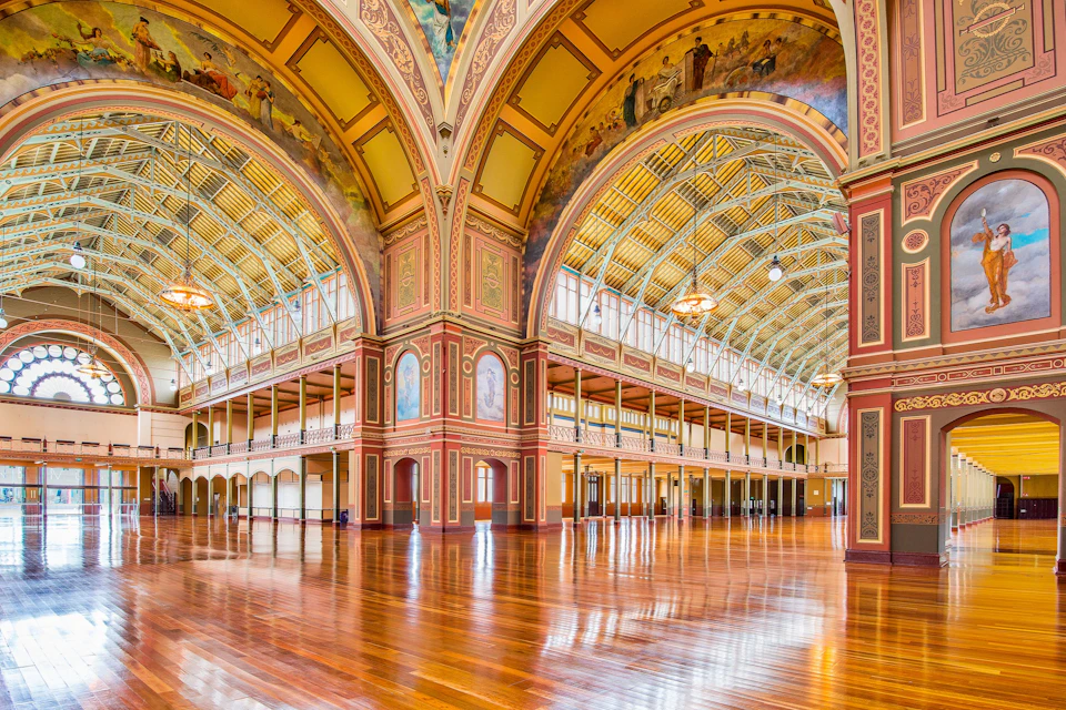 A grand, empty hall with polished wooden floors, ornate arches, murals on walls, and a high, decorated ceiling with chandeliers.