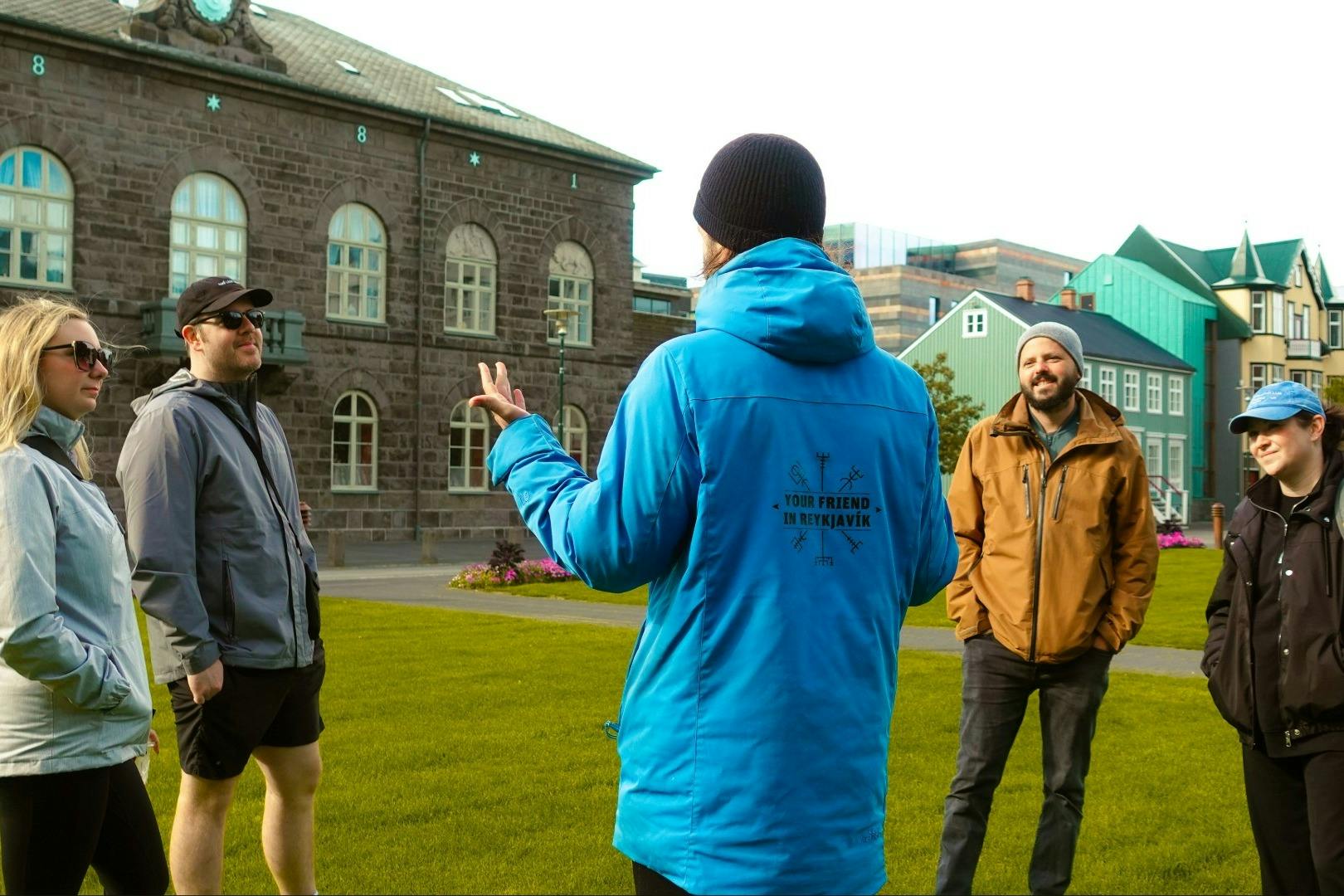 파란색 재킷을 입은 투어 가이드가 공원에서 화려한 건물을 배경으로 두 사람에게 말을 건넨다.