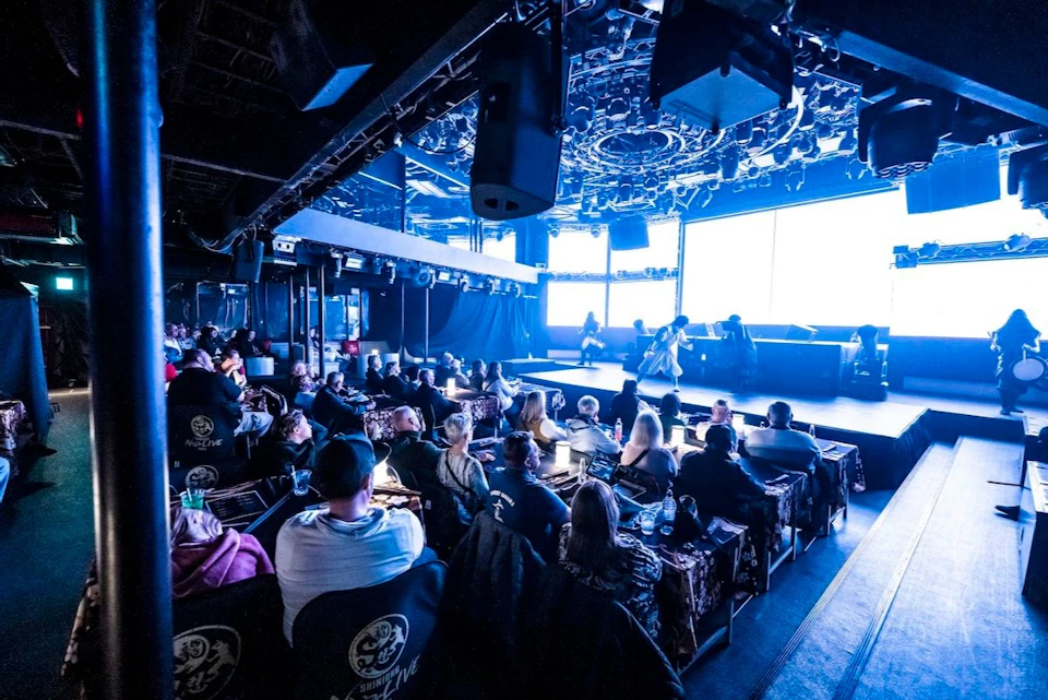 A live performance venue with seated audience, a stage, and performers under colorful lighting.