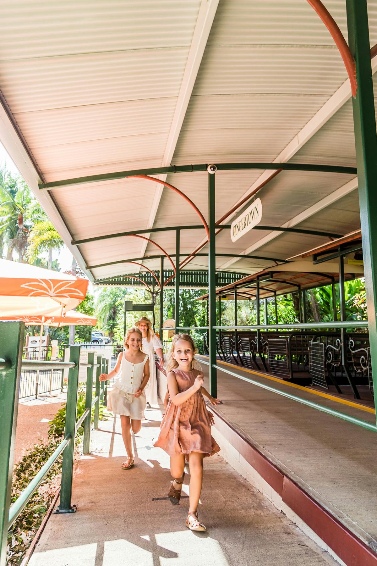 三个年轻女孩在阴凉的人行道上愉快地跑步，旁边有一个写着“Endertom”的标志。 背景包括绿色植物和空长椅。