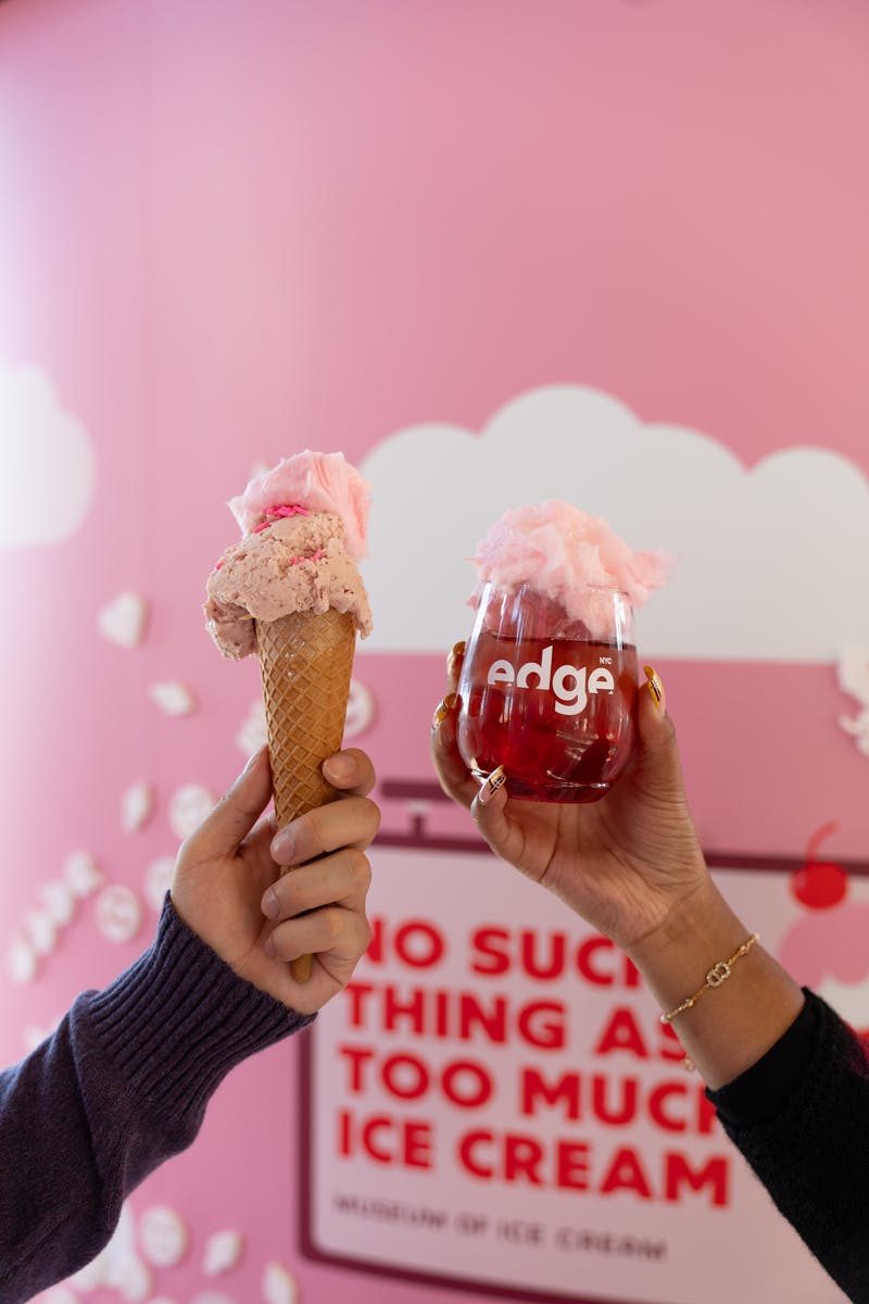 Dvě ruce držící růžový kornout zmrzliny a sklenici červeného nápoje s cukrovou vatou na růžovém pozadí.