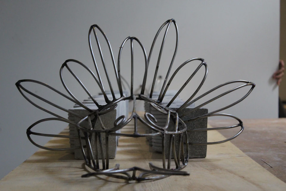 Wire sculpture resembling a crown with petal-like loops, set against a neutral background. Concrete blocks support the structure.