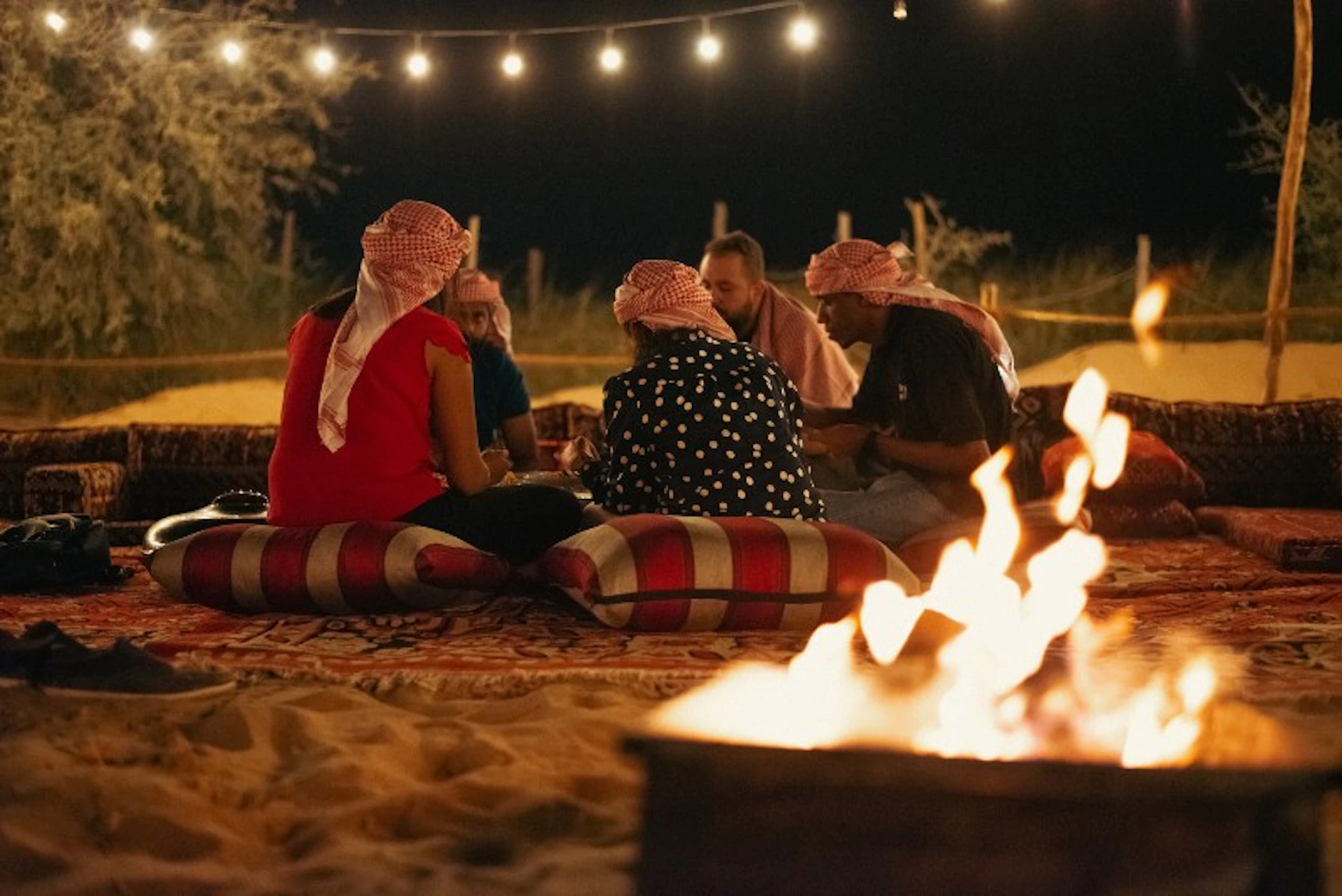 A group of people seated on cushions around a fire at night, wearing traditional headscarves, with string lights overhead.