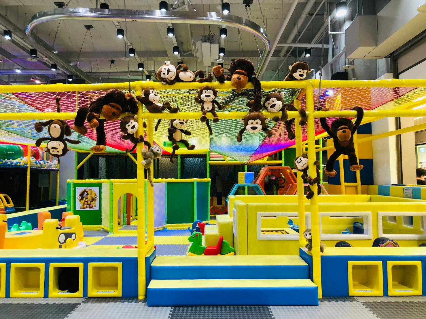Bright indoor children's play area with a colorful climbing net, hanging monkey toys, and soft structures for play.