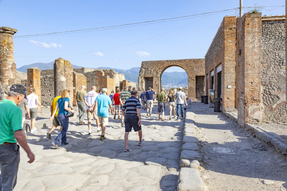 A group of tourists walks through the ancient ruins of Pompeii under a clear blue sky.