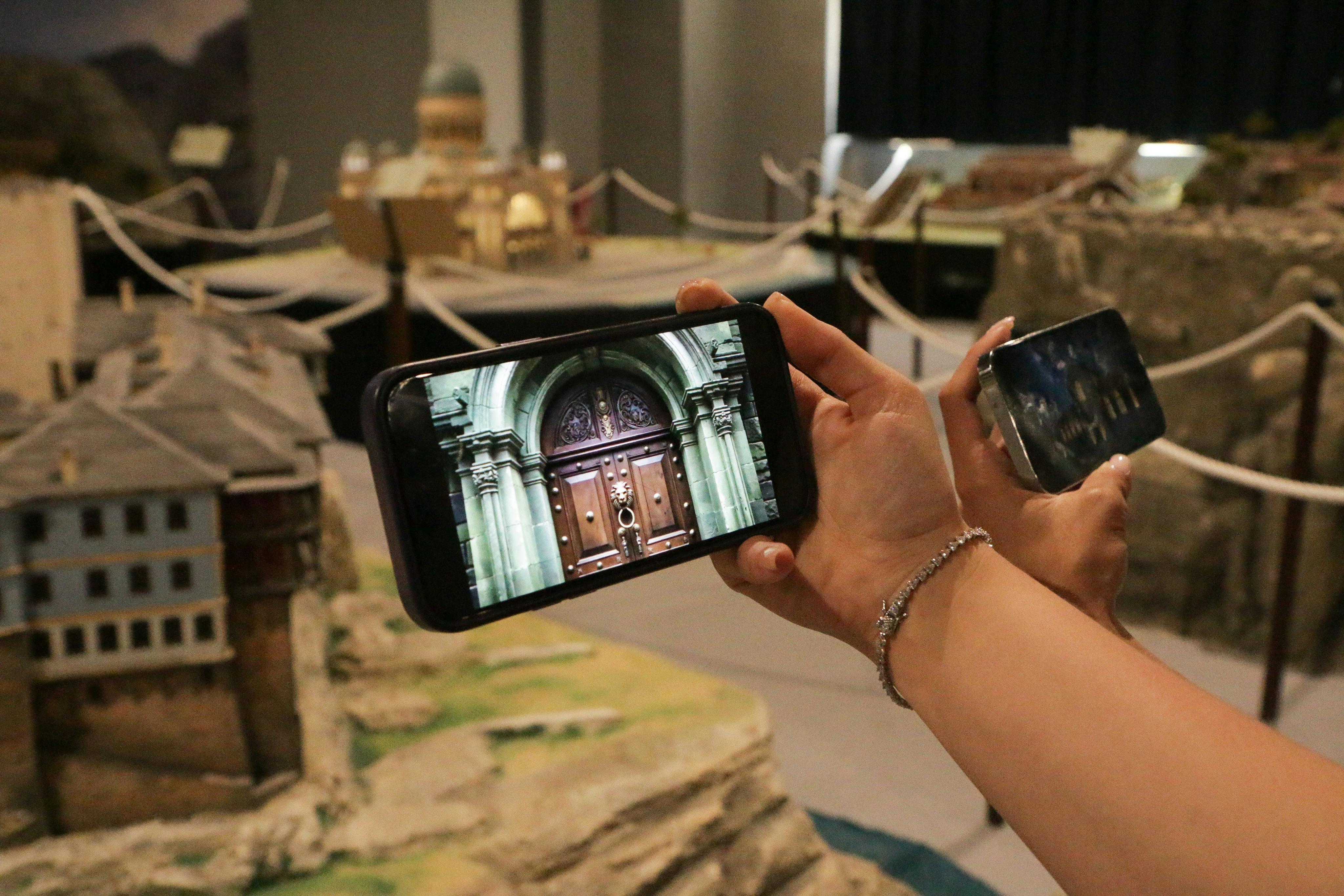Two people hold smartphones capturing images of architectural models, focusing on a detailed model of a building facade.