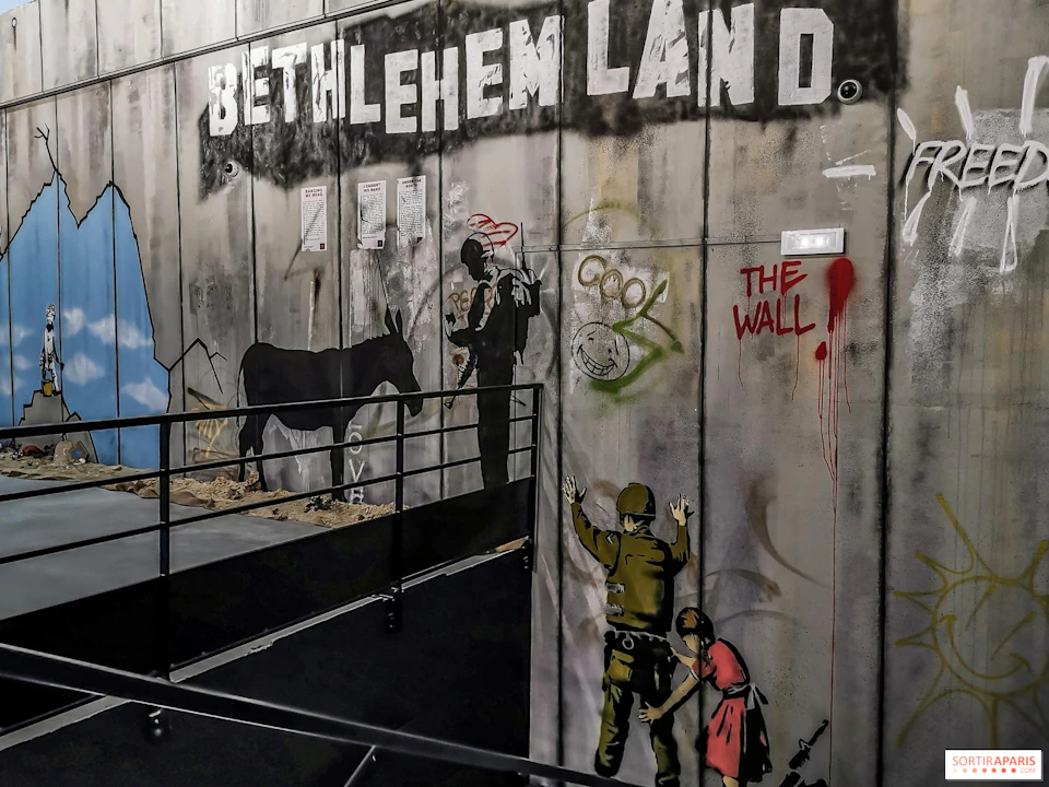 Graffiti on a wall shows soldiers, a donkey, and children. Text reads "BETHLEHEM LAND" and "THE WALL" in large letters.