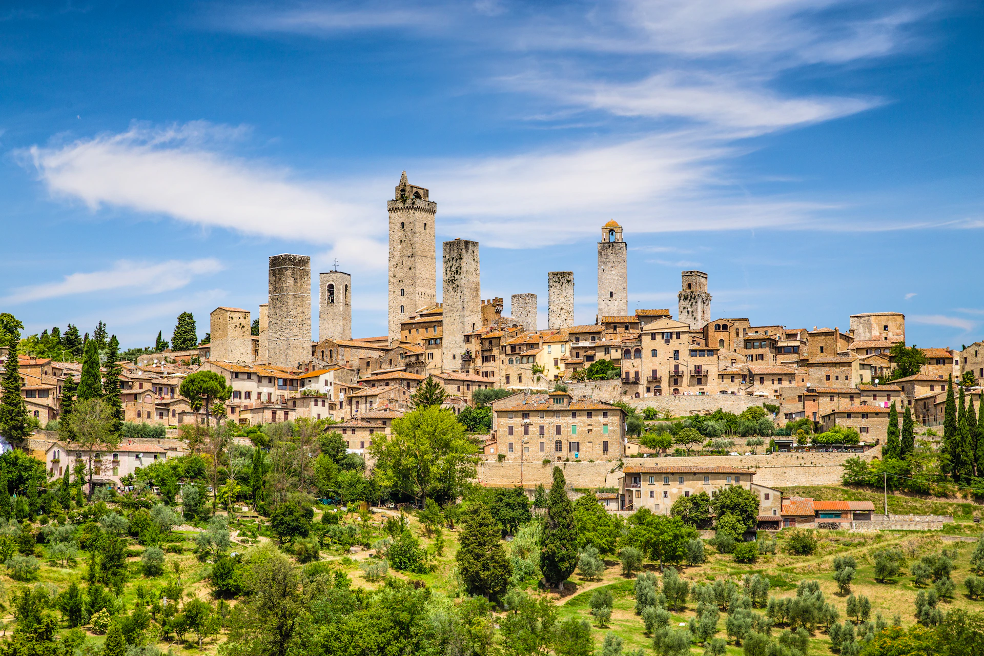 A historic town with numerous tall stone towers, surrounded by lush greenery under a blue sky with scattered clouds.