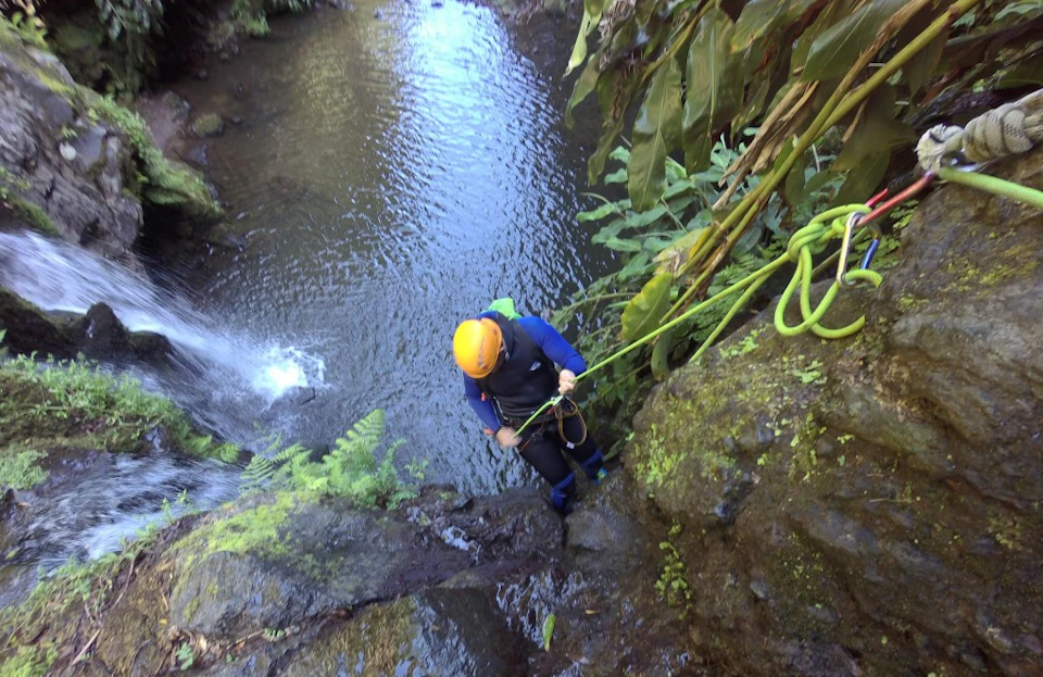 A person in a wetsuit and helmet rappelling down a steep, mossy rock near a waterfall and a flowing river.