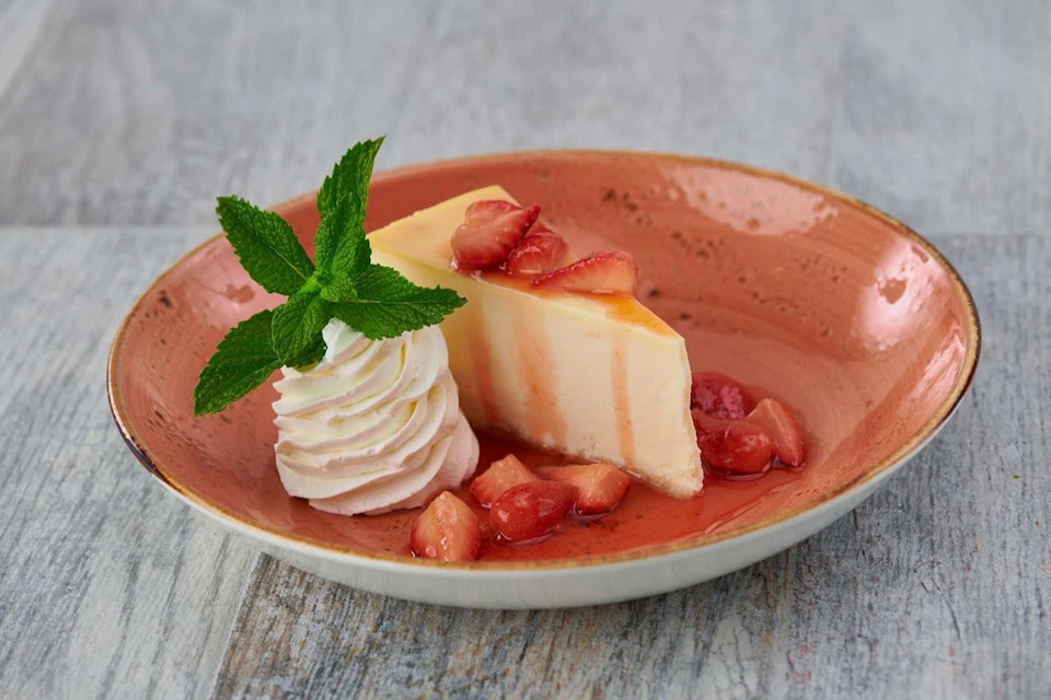 A slice of cheesecake with strawberry topping, garnished with whipped cream and mint, served on an orange plate.