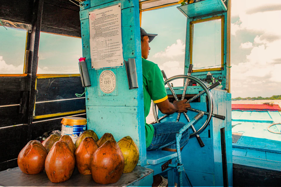 A person in a green shirt steers a boat with coconuts and containers nearby, against a backdrop of sky and water.