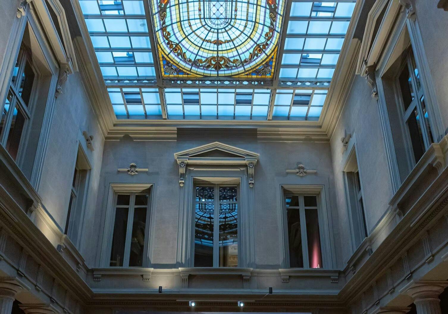 An interior space with a decorative stained glass ceiling and classical architectural details surrounding tall windows.