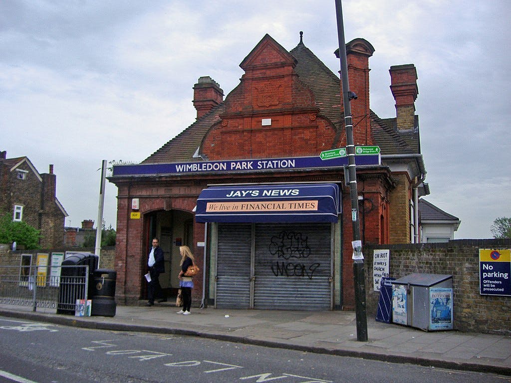 Un edificio de ladrillo rojo rotulado Estación de Wimbledon Park, con un quiosco de prensa cerrado y dos personas caminando fuera.