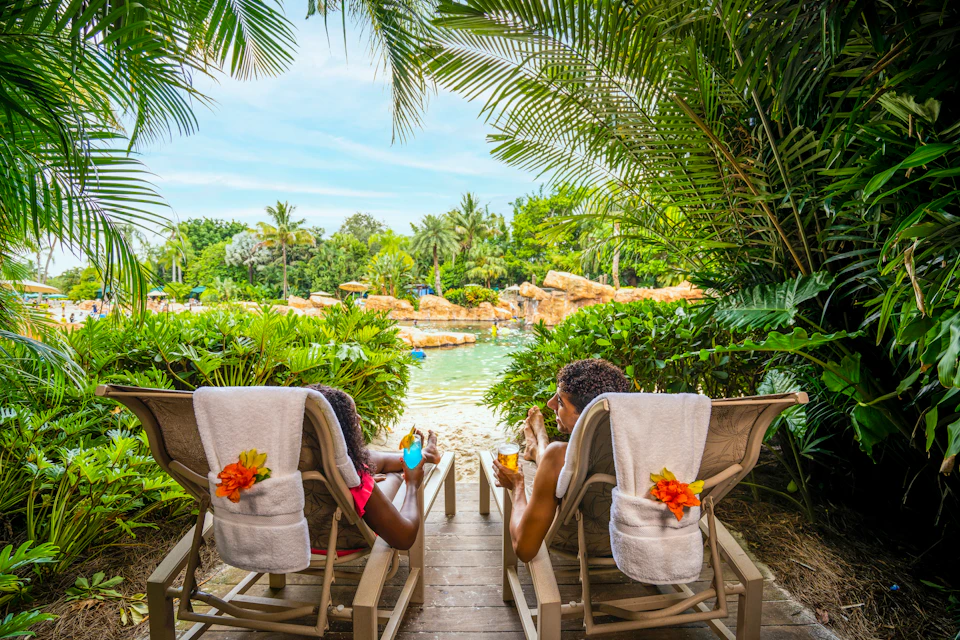 Two people lounge on chairs with drinks, surrounded by tropical plants, facing a lush, green view with rocks and water.