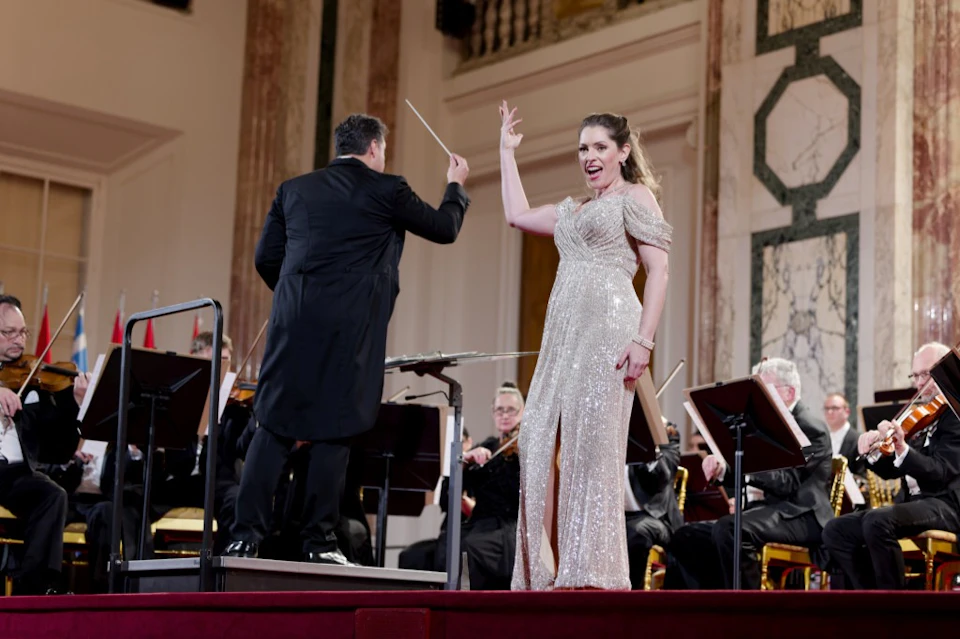 A conductor leads an orchestra as a performer in a sparkly gown sings on stage in an elegant concert hall.