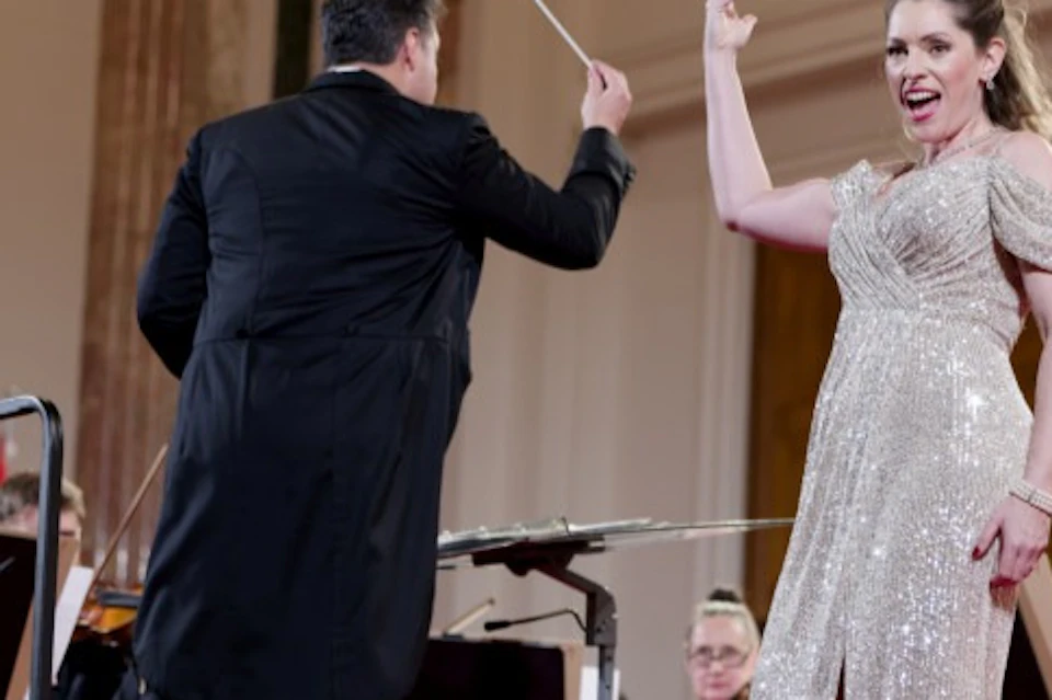 A conductor leads an orchestra as a performer in a sparkly gown sings on stage in an elegant concert hall.