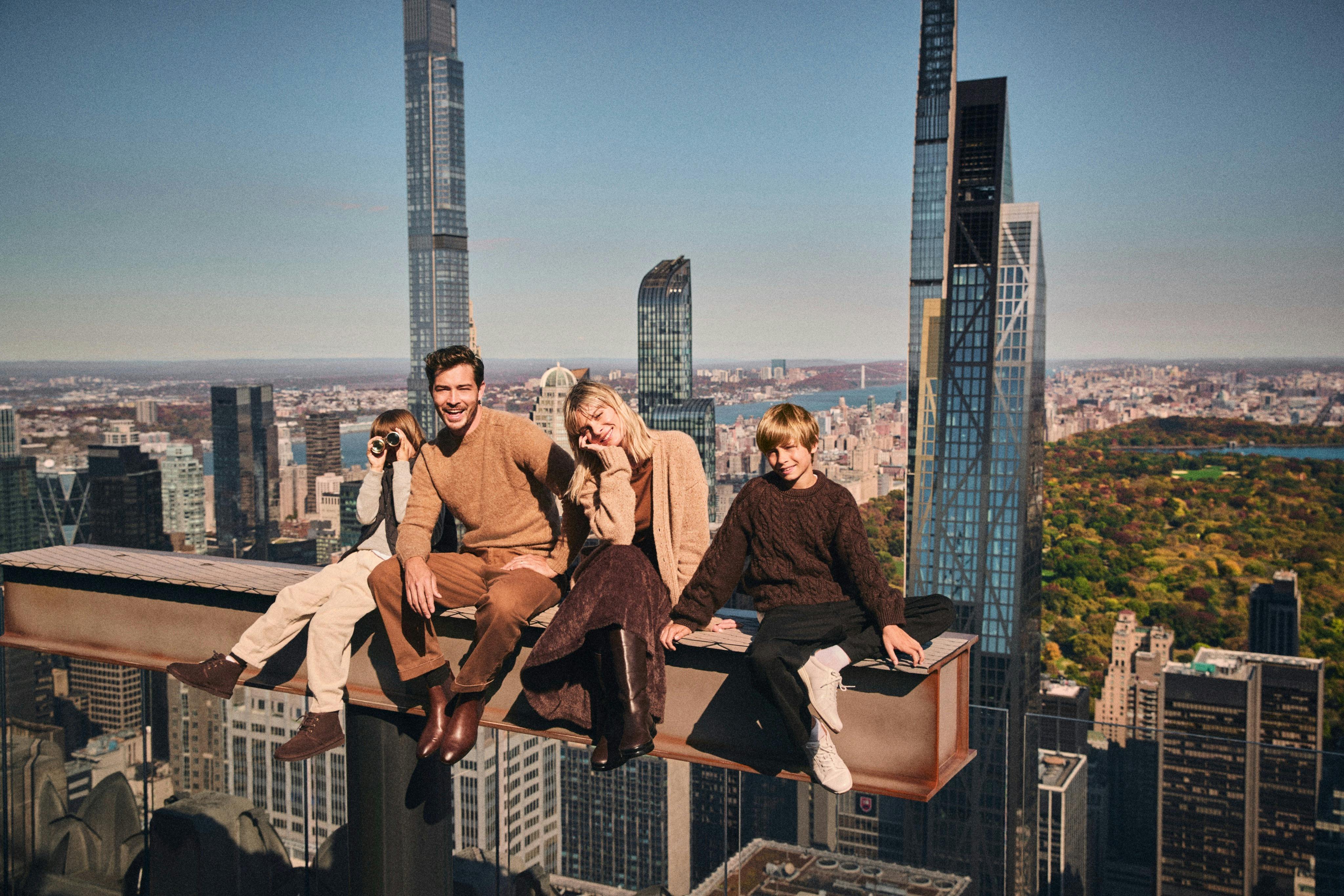 Four people sitting on a steel beam above a cityscape, wearing fall clothing, with tall buildings in the background.