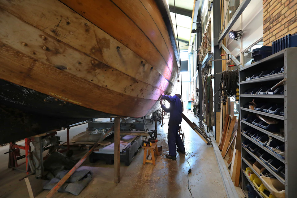 A person working on the wooden hull of a boat in a workshop, surrounded by tools and equipment.