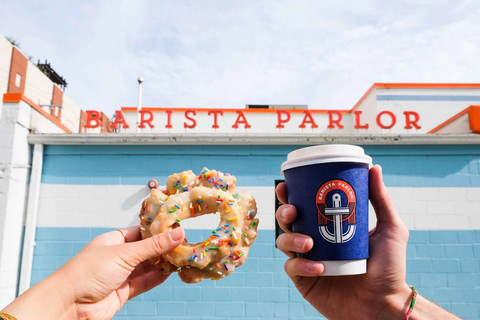 Two hands holding glazed donuts with sprinkles and a blue coffee cup in front of a building with "Barista Parlor" sign.