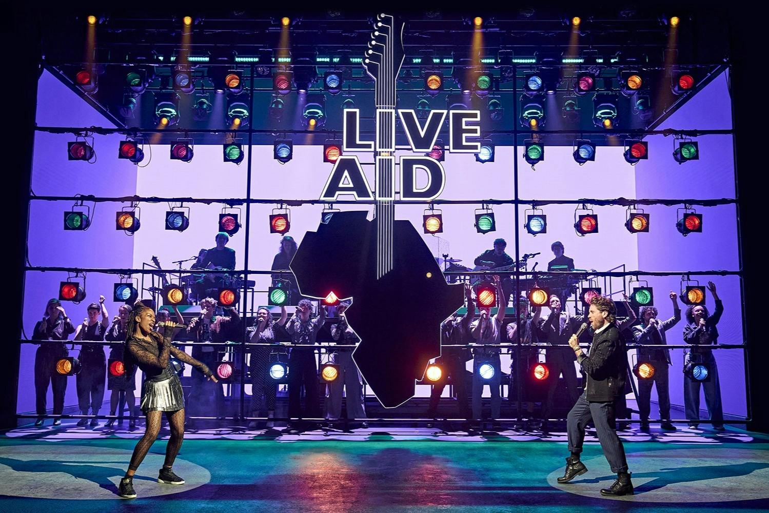 一位表演者在舞台上演唱“LIVE AID”标志，五颜六色的灯光和乐队在他身后演奏，观众欢呼。