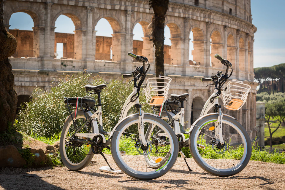 Five white bicycles parked near green bushes with the Colosseum in the background on a sunny day.